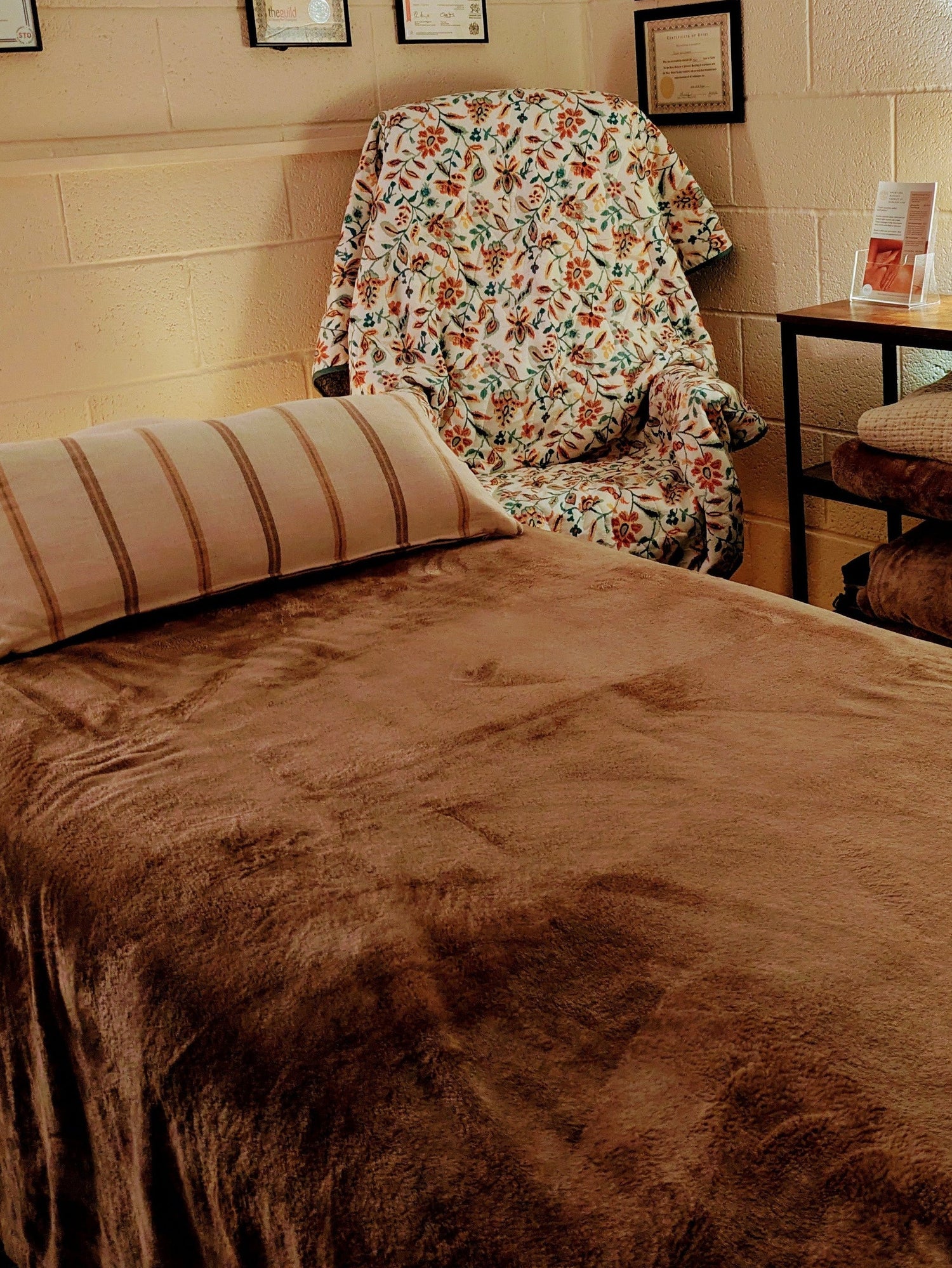 Treatment room with a massage table covered in brown sheets, floral chair, and framed pictures on the wall.